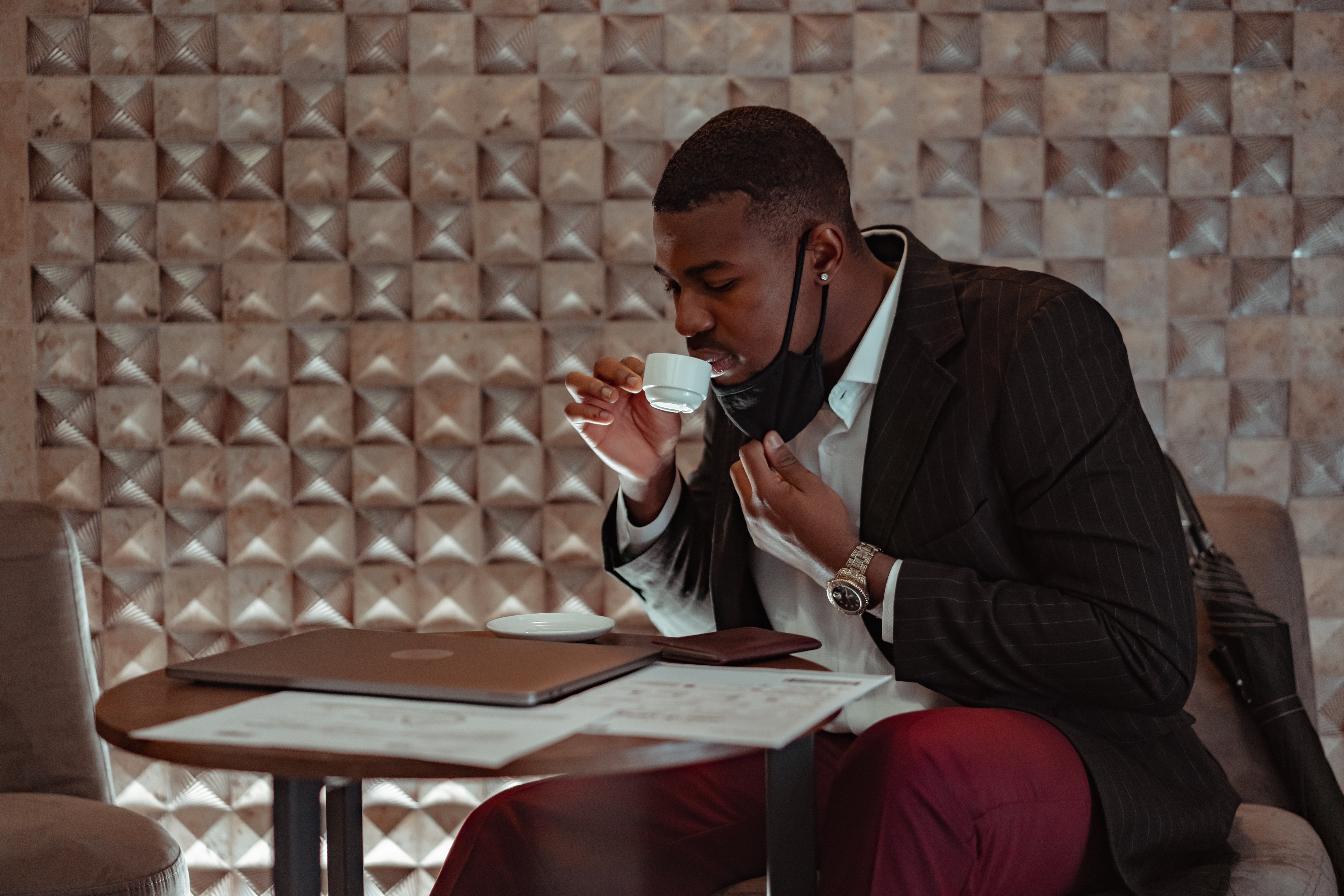 A man sipping coffee from a small cup while sitting down wearing black blazer